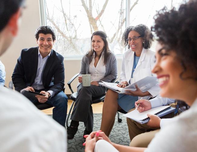 A group of doctors sit in a circle indoors, smiling and speaking to each other. They are dressed either in white coats or in business attire, and some hold notebooks, binders, or tablets in their hands. 