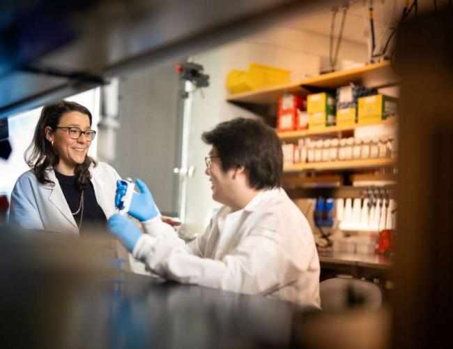 Dr. Cristina Puig-Saus is pictured wearing a white lab coat while standing in a laboratory and speaking with another scientist. Both individuals are smiling. 