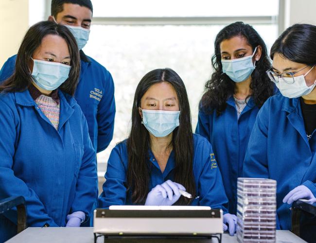 Dr. Melody Li, centered, gestures to a benchtop instrument while four masked researchers in blue lab coats watch closely. A stack of clear culture plates sits on the lab bench beside them.