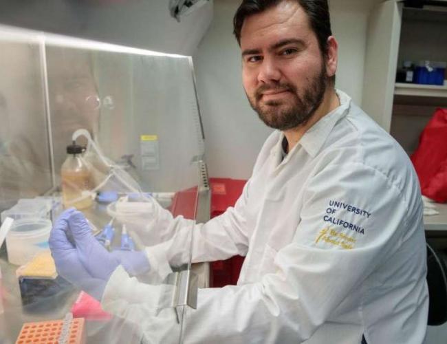 Gustavo Garcia Jr. sits with gloved hands beneath a UCLA lab hood.