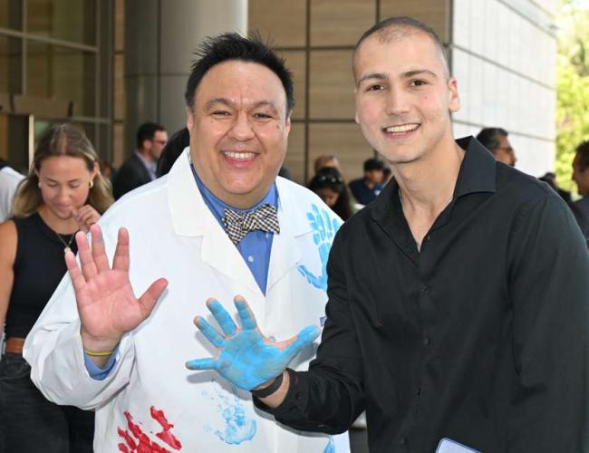 a photo with pediatric cancer researcher Dr. Steve Jonas wearing a white coat on the left posing with cancer survivor Sina Sinbari on the right wearing a black collared long sleeve shirt at UCLA Mattel Children's Hospital