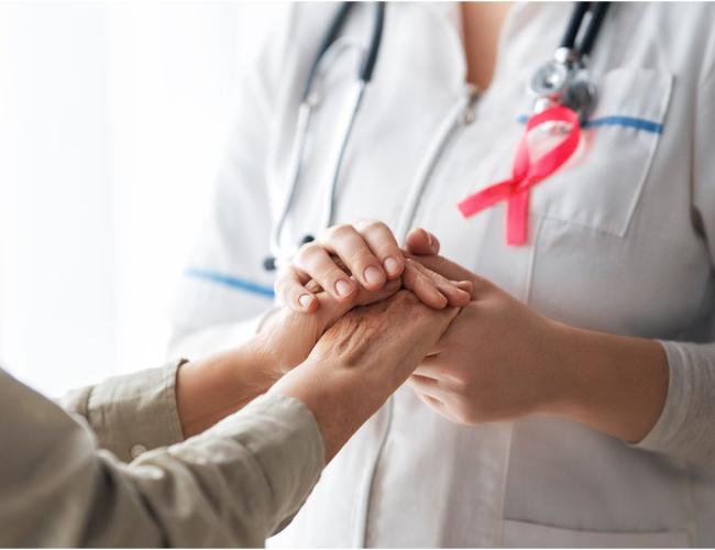 A patient, left, and medical care provider holding hands; provider has a pink breast cancer ribbon on her lapel