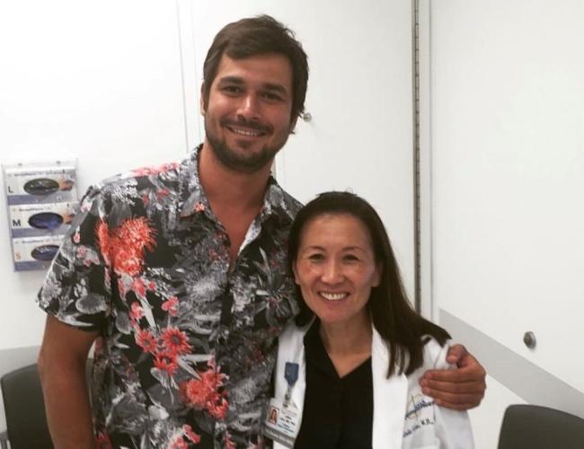 Attorney and brain cancer survivor Jamil Newirth stands smiling beside Dr. Linda Liau, chair of neurosurgery at the David Geffen School of Medicine at UCLA and director of the UCLA Brain Tumor Program, in a medical office setting. Newirth wears a colorful floral shirt while Dr. Liau wears a white medical coat.