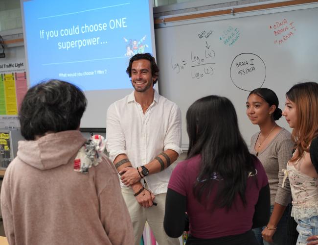 A man smiles while talking with a small group of students in a classroom, standing in front of a whiteboard and a projected slide that reads "If you could choose ONE superpower... What would you choose? Why?"
