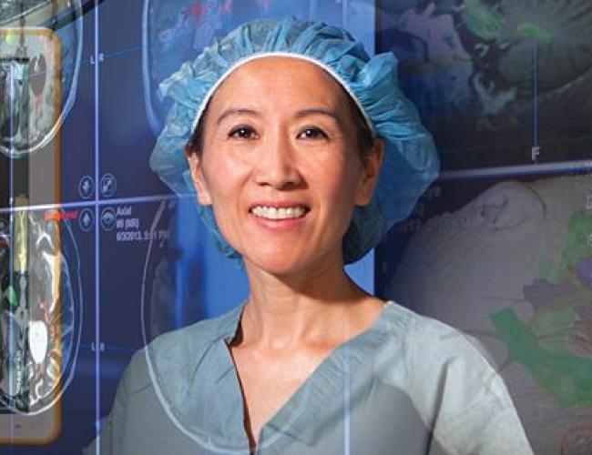 Dr. Linda Liau in surgical scrubs and cap standing in an operating room with brain scan images and surgical lighting visible in the background.