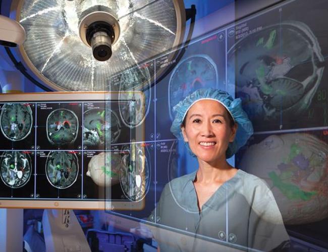 Dr. Linda Liau in surgical scrubs and cap standing in an operating room with brain scan images and surgical lighting visible in the background.