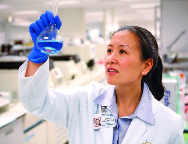 Dr. Linda Liau in a white lab coat and blue gloves examining a petri dish in a laboratory.