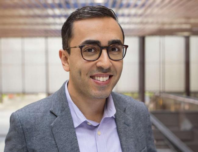 Mina Sedrak Smiles for a photograph. He wears grey and purple.