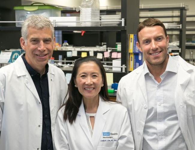 Thomas Graeber, Dr. Linda Liau, and David Nathanson standing together in white lab coats in a laboratory setting, smiling at the camera.