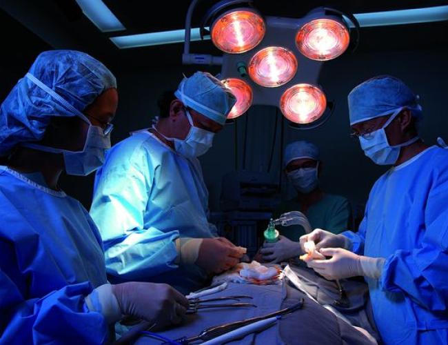 A group of surgeons gather around a patient in an operating room. The lights are dimmed and blue-tinged and overhead light blares above.