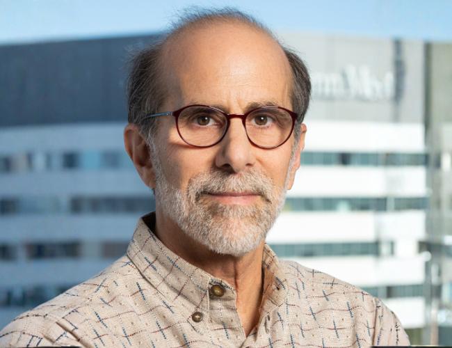 Headshot of Kenneth Zaret in a tan button up and glasses against a cityscape