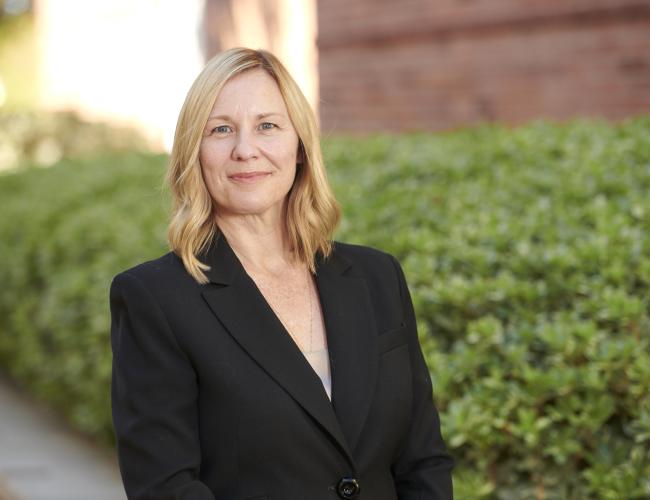 Photograph of Amander Clark, PhD wearing a black suit jacket and standing in front of greenery. 