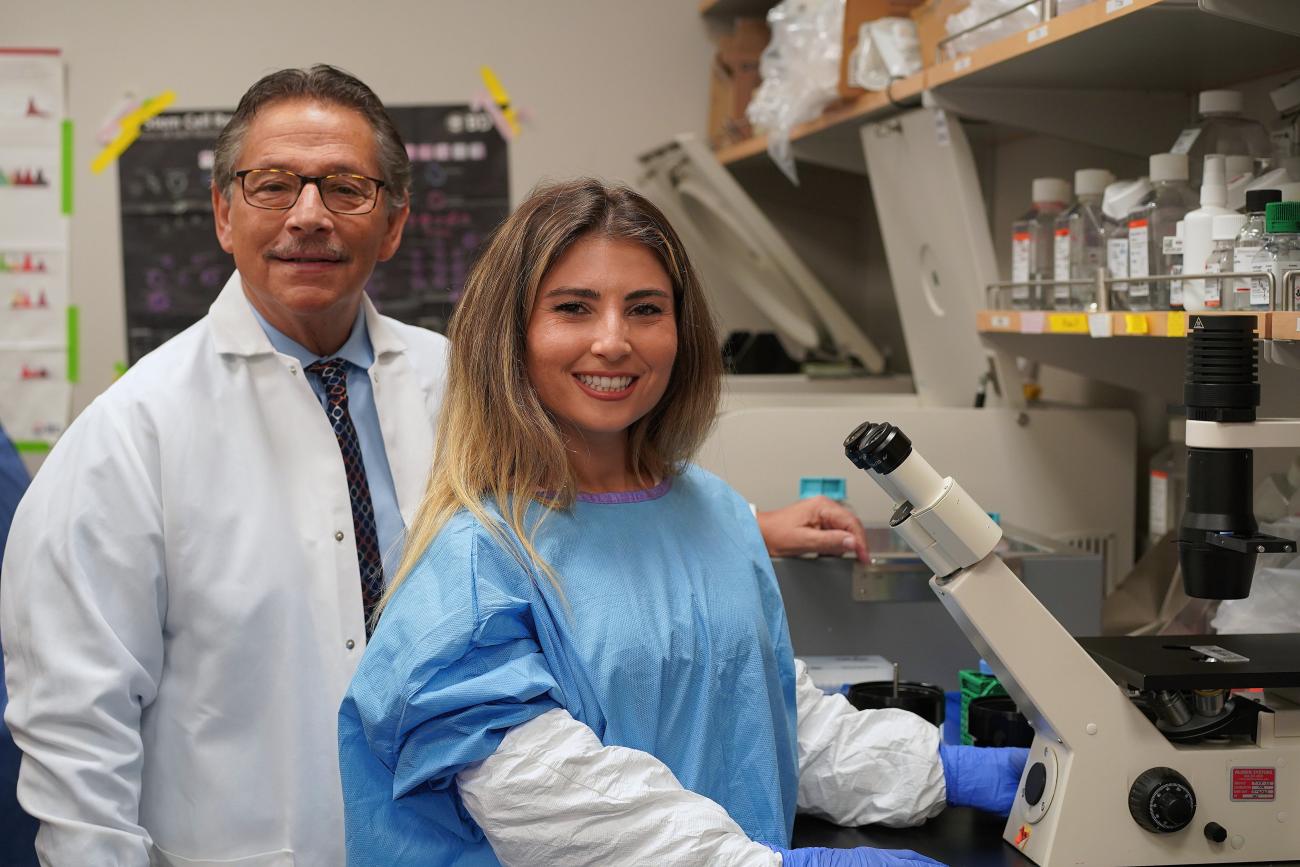 Scientist Donald Kohn on the left wearing a white lab coat and scientist Eva Segura on the right wearing lab protective gear; both are in the lab next to a microscope