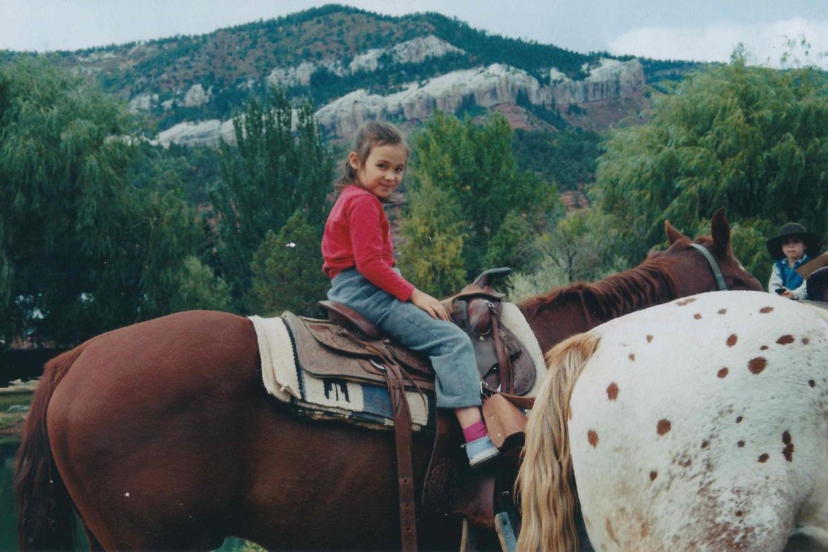 Young child in red sweater riding a spotted horse in a mountainous landscape