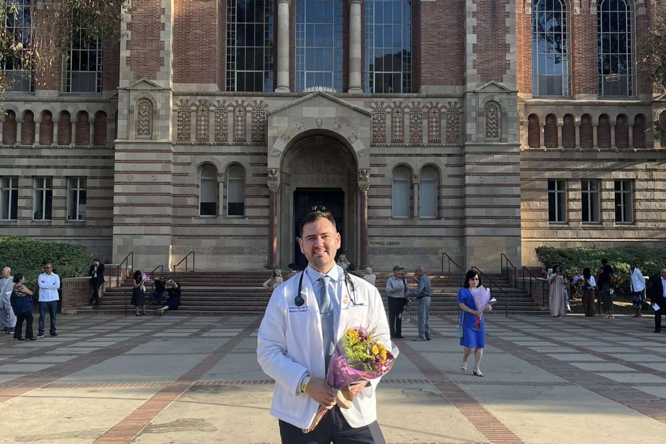 First-year medical student Gustavo Garcia Jr. wears a lab coat, a stethoscope over his shoulders, and holds a bouquet of flowers as he smiles for a photo outdoors.