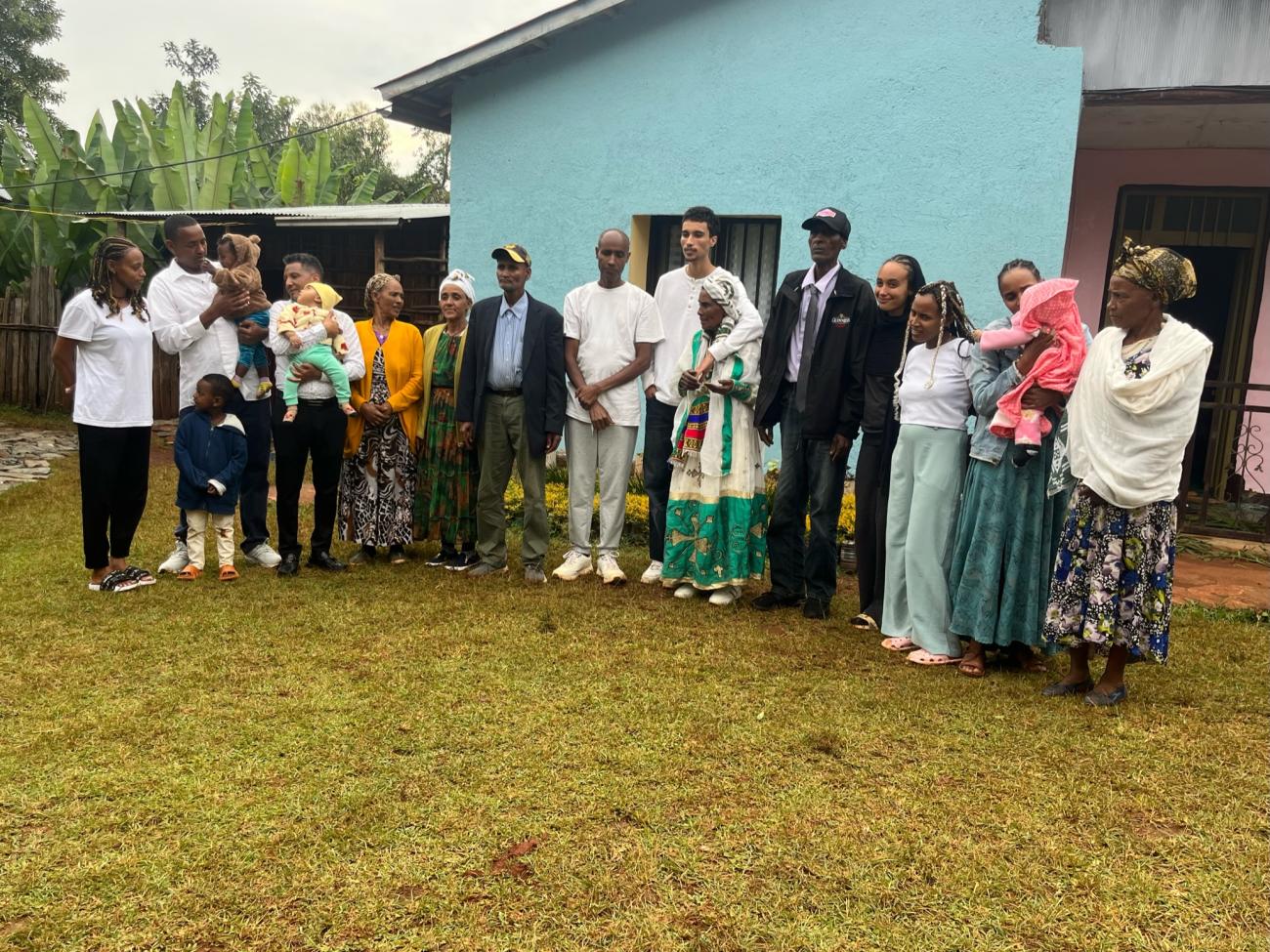 A multigenerational group of about a dozen people, including young children and babies, pose together outdoors in front of a light blue house with lush green vegetation in the background. Several people wear traditional Ethiopian clothing, including colorful embroidered garments and head wraps, while others are dressed in casual or formal Western attire.