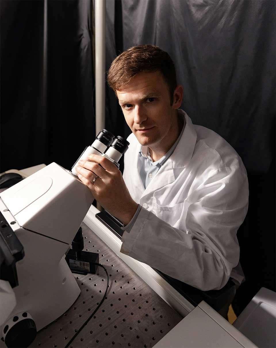 Aaron Meyer in white lab coat using microscope in laboratory setting with dramatic lighting.