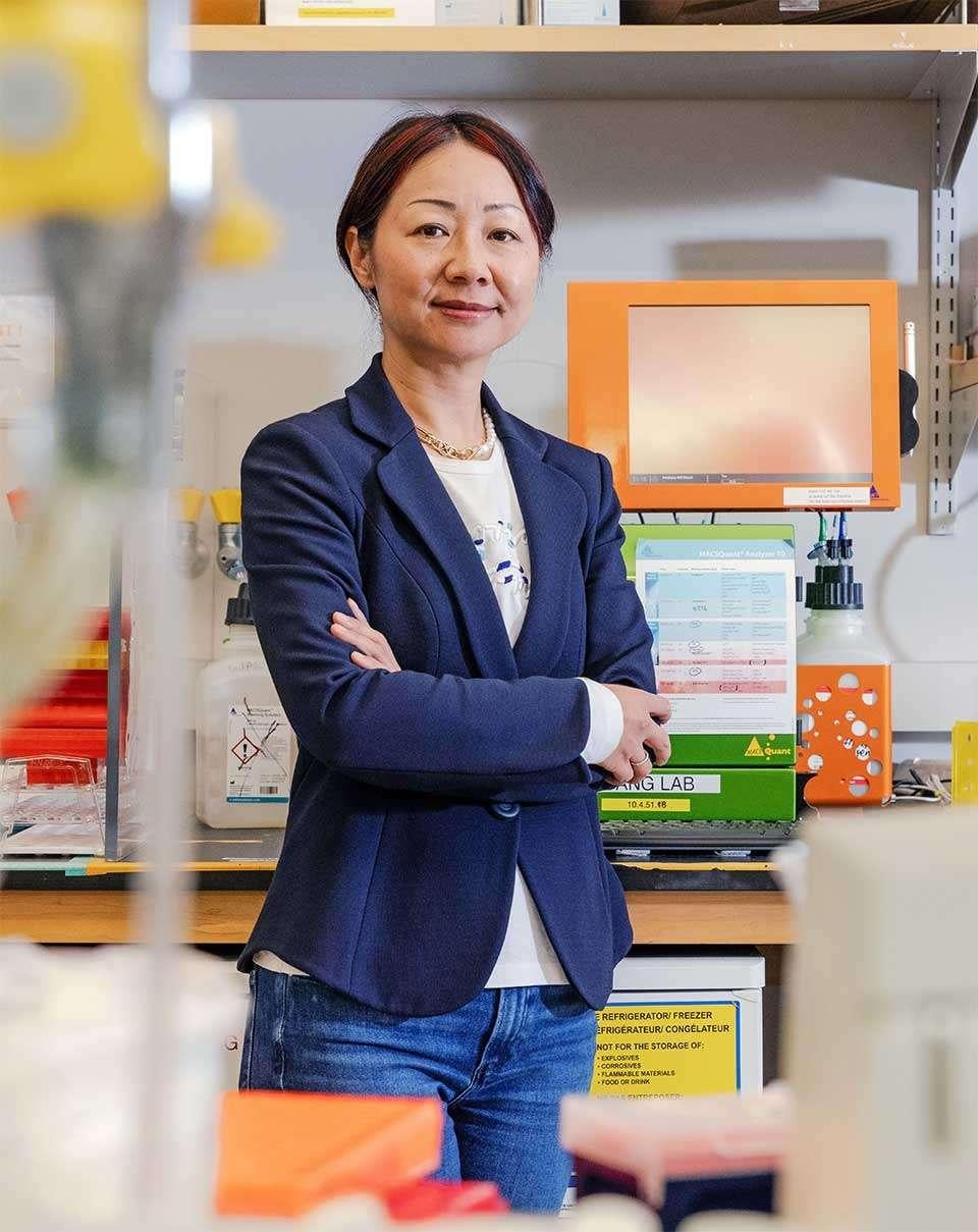 Dr. Lili Yang in navy blazer with arms crossed standing in laboratory setting with colorful storage boxes and equipment.