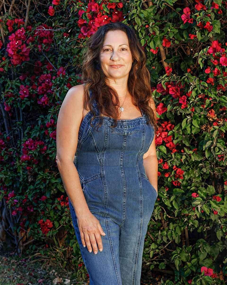 Dr. Memarzadeh in denim dress standing in front of vibrant red and pink bougainvillea flowers in garden setting.