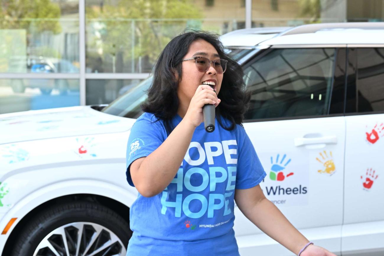 Pediatric cancer survivor Angela Unayan wearing a blue t-shirt that says HOPE holding a microphone to speak at the Hyundai Hope on Wheels ceremony