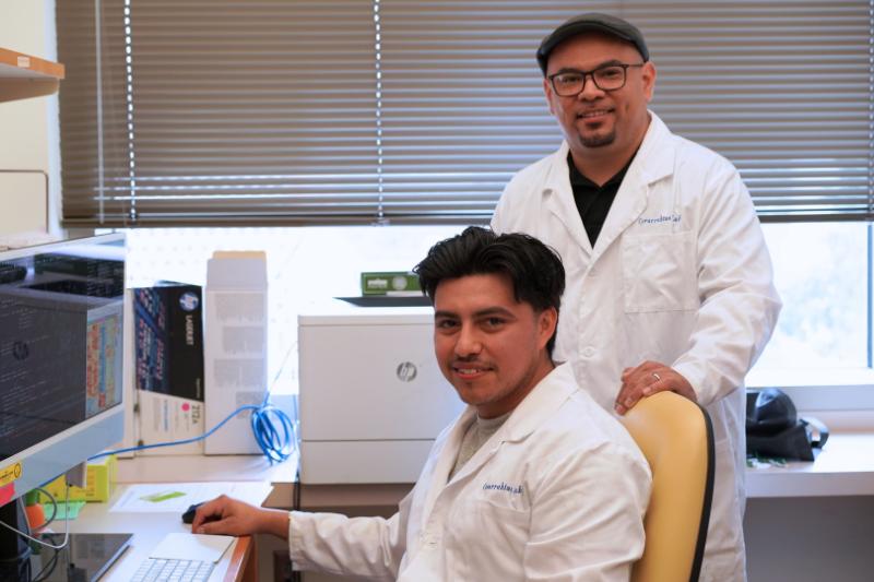 Two researchers wearing white coats smile for a photo in a UCLA lab: Ivan Salladay-Perez (left) and Anthony Covarrubias (right).