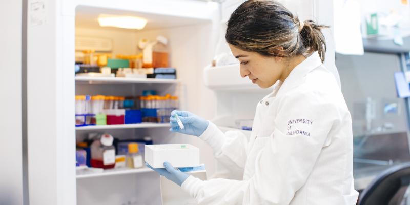 A scientist looks at a vial of liquid in front of a refrigerator in a lab