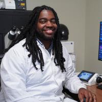 Headshot of Dr. D'Juan Farmer wearing a white lab coat sitting next to a computer monitor with zebrafish on the screen