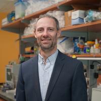 UCLA scientist Peter Clark smiles in his lab wearing a checkered shirt and dark grey suit jacket.