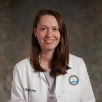 UCLA physician-scientist Jennifer Chia smiles for a headshot in a white labcoat in front of a muted brown background.