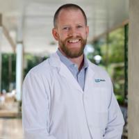 UCLA researcher Vondriska, Thomas smiles for a photograph. He wears a white UCLA labcoat and stands indoors with a green landscape appearing behind him.