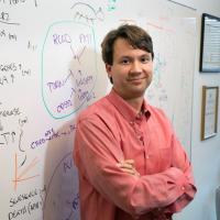 William Lowry stands smiling with crossed arms in his office before a whiteboard.
