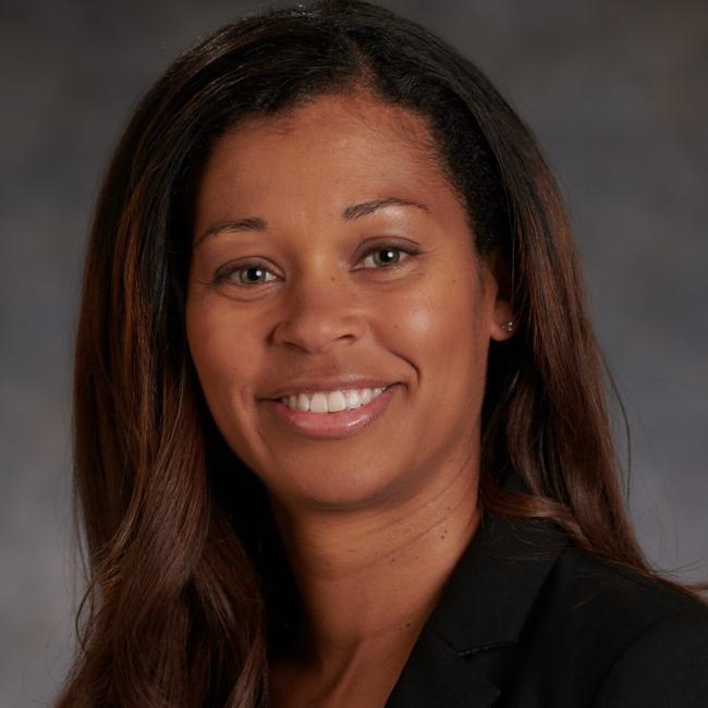 A headshot of Janai Carr-Ascher smiling, with dark hair, wearing a black blazer against a grey background.