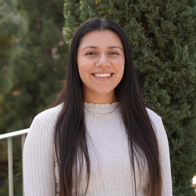 Headshot of BSCRC trainee Jessenya Mil, a woman with long straight dark hair wearing a white ribbed top, smiling outdoors.
