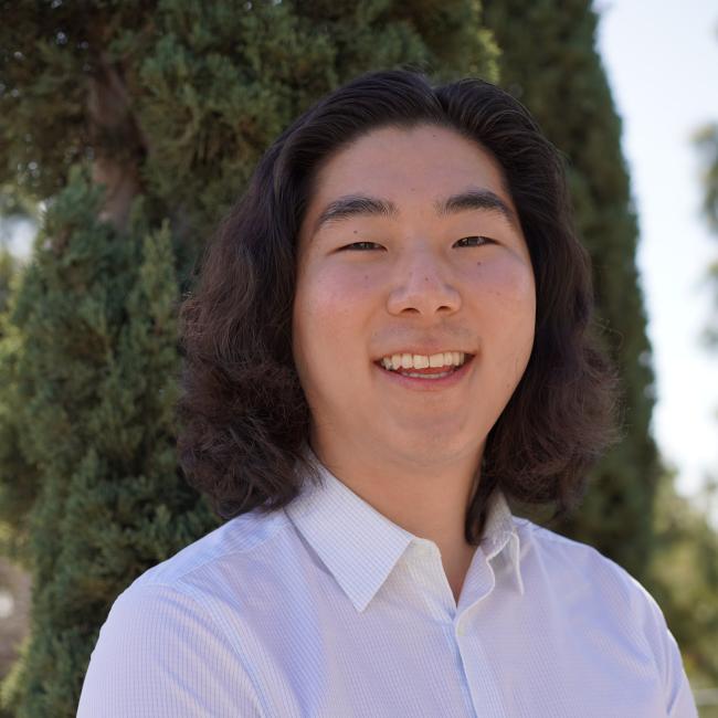 Headshot of Sang Pil Yoo (Alex), a man with shoulder-length wavy dark hair wearing a light-colored button-up shirt, smiling outdoors.