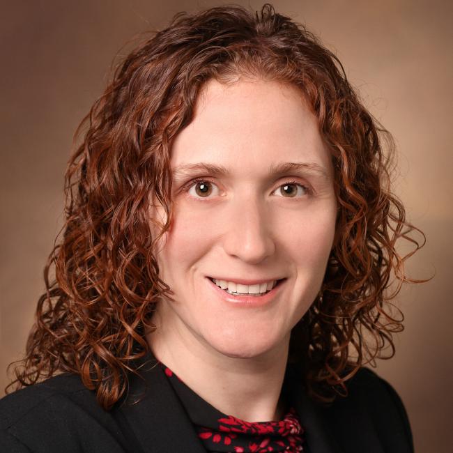 Headshot of Jennifer Sucre, a person with curly reddish-brown hair, smiling against a neutral background, wearing a black blazer and patterned top.