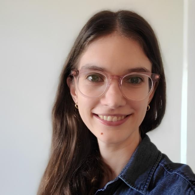 Headshot of BSCRC trainee Grace Hancock, an individual with long dark hair and clear-framed glasses wearing a denim collared shirt, smiling indoors against a white background.