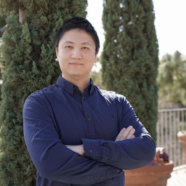 Headshot of Xuegang Yuan, an individual with short dark hair wearing a dark blue button-up shirt, posing with arms crossed outdoors.