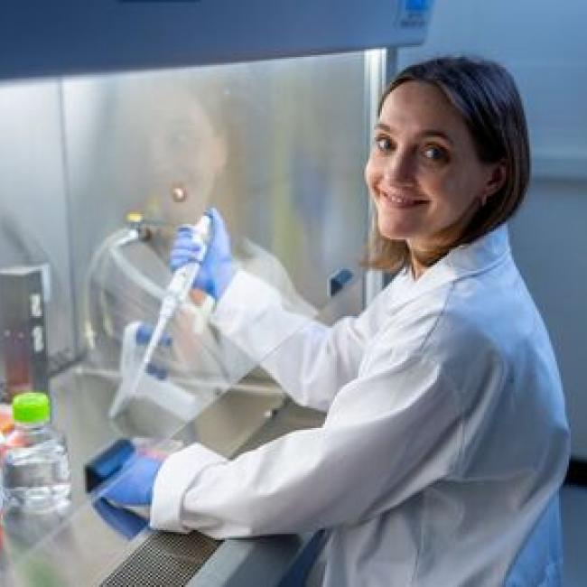 Katrina Adams, wearing a white lab coat and blue gloves, smiling while working at a biosafety hood in a laboratory setting.