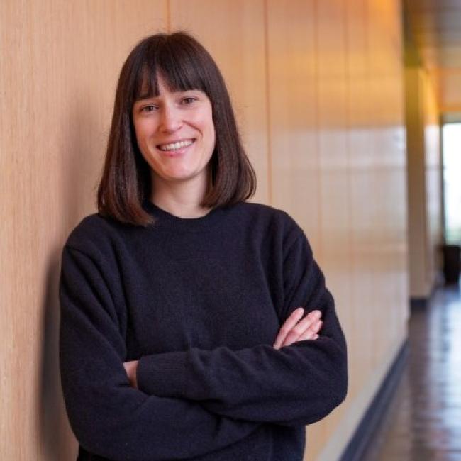 Headshot of Julia Aguadé Gorgorió, a person with shoulder-length dark hair with bangs, smiling with arms crossed in an indoor hallway setting, wearing a black sweater.