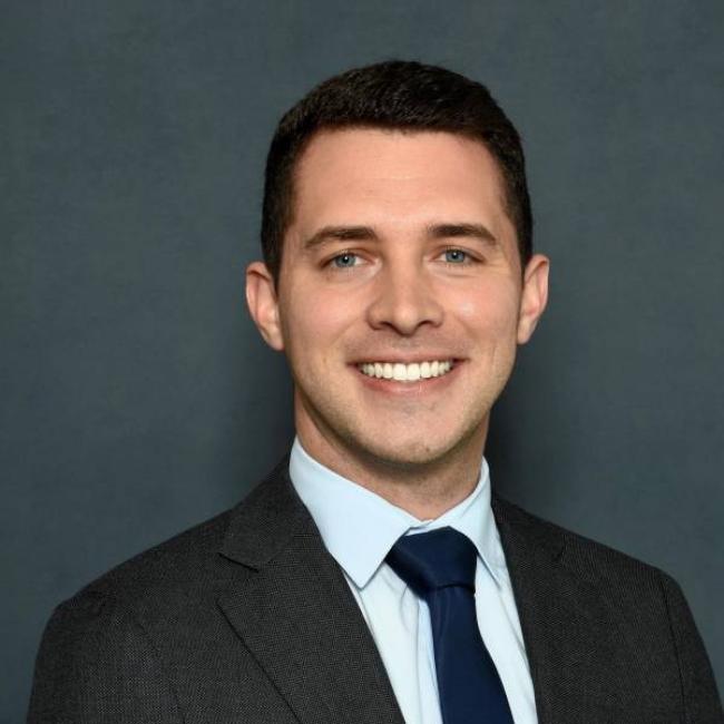 Headshot of Cody Aros, a person with short dark hair and blue eyes, smiling against a dark gray background, wearing a gray suit jacket, light blue shirt, and navy tie.
