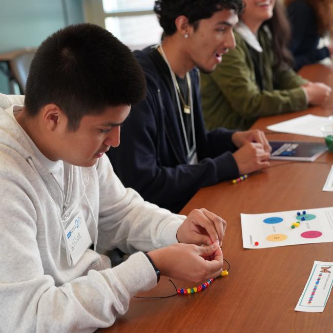 a male high school student is making a bracelet seated next to another male high school student