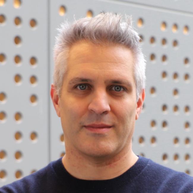 Headshot of Vincenzo Calvanese, a person with short gray hair and blue eyes, against a perforated metal wall background, wearing a dark navy sweater.