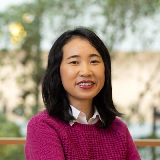 Headshot of Julia Chin, a person with shoulder-length dark hair, smiling against a blurred outdoor background, wearing a pink knit sweater.