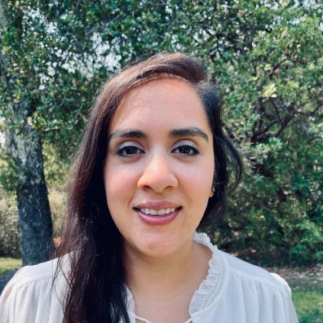 Headshot of Akanksha Chhabra, a person with long dark hair, smiling outdoors against a blurred green tree background, wearing a white top.