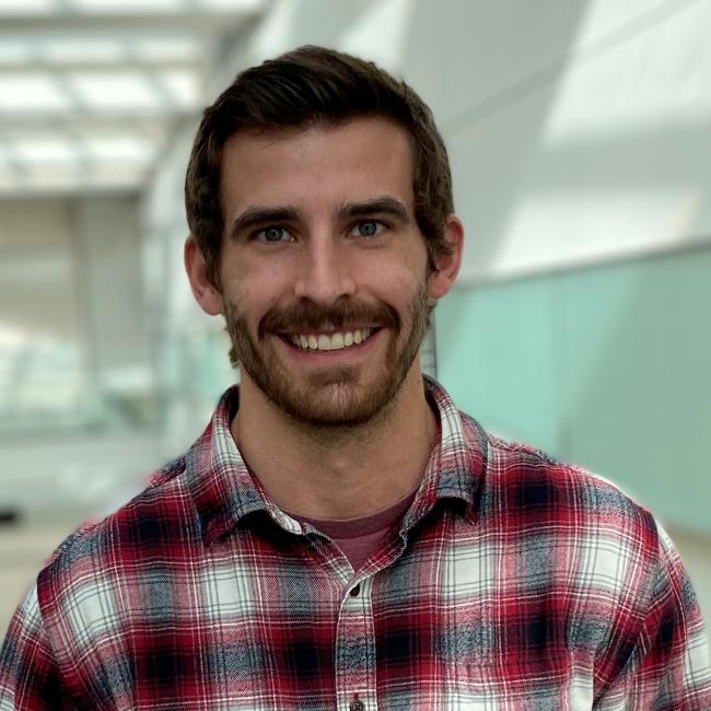A headshot of Dan Jaklic dressed in a red, white, and black plaid button-up shirt. He is smiling and has dark hair.