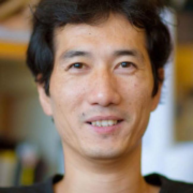 Headshot of Di Chen, a man with short black hair, smiling in an indoor setting with a warm, blurred background.