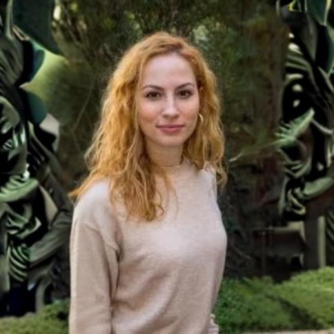 Headshot of BSCRC trainee Elena Korsakova, an individual with long wavy blonde hair wearing a light beige sweater, standing outdoors against a green foliage background.
