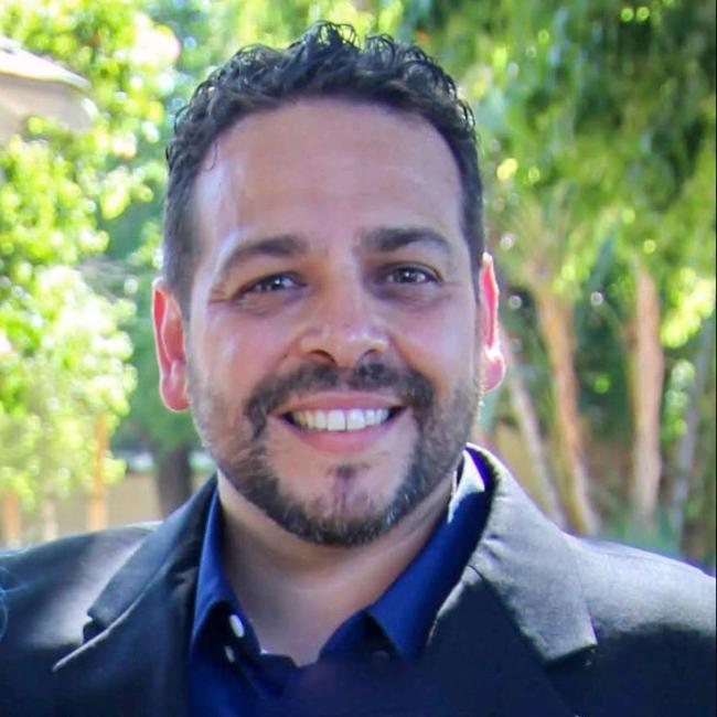 Headshot of Enrique Sosa, a man with short dark curly hair and a beard, smiling outdoors against a blurred green background, wearing a blue shirt and blazer.