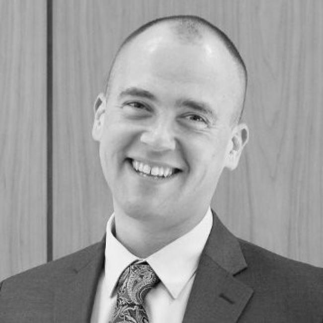 Headshot of Eric Gschweng, a person with a shaved head, smiling against a wooden background, wearing a suit jacket and patterned tie. Black and white photo.