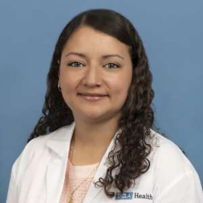 Headshot of Vivian Fajardo, a person with long dark curly hair, smiling against a blue background, wearing a white lab coat with a UCLA Health logo.
